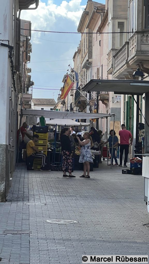 Schmaler Straßenzug mit kleinen Marktständen in Petra auf Mallorca, Menschen unterhalten sich entspannt vor frischem Obst und Gemüse