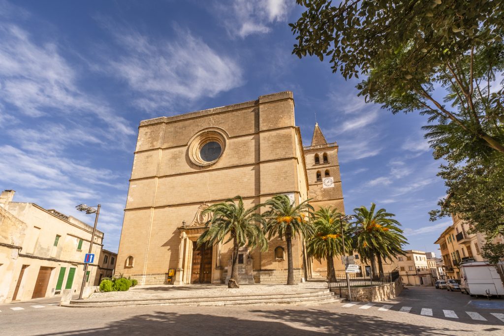 Frontansicht der Pfarrkirche Nostra Senyora de la Consolació in Porreres, Mallorca, mit Palmen, blauen Himmel und historischem Uhrenturm