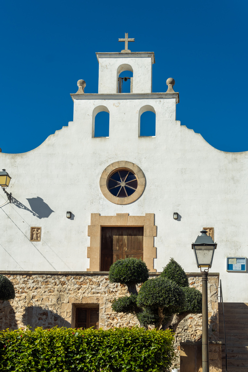 Katholische Kirche in Portopetro mit weißer Fassade, Rundfenster und Glockengiebel vor blauem Himmel.