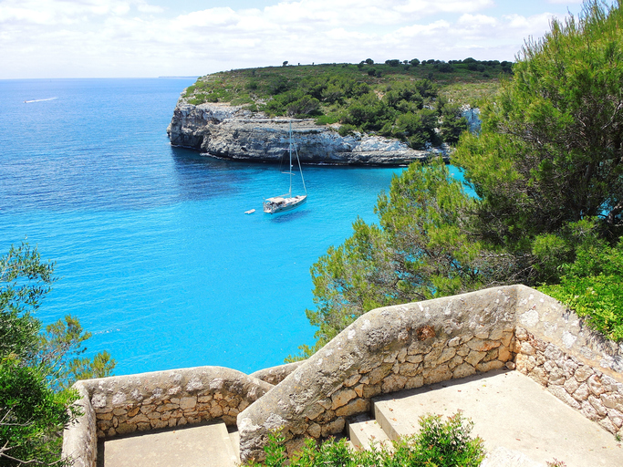 Steintreppe mit Ausblick auf die Bucht Cala Romàntica und ein vor Anker liegendes Segelboot im klaren, blauen Mittelmeerwasser.