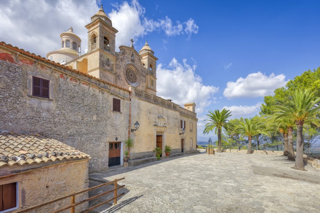 Historisches Kloster Ermita de la Mare de Déu de Bonany nahe Petra auf Mallorca mit Sandsteinfassade, Rosettenfenster, Türmen und Palmenallee vor blauem Himmel.