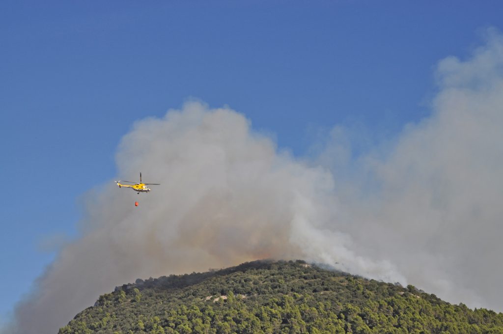 Ein gelber Löschhubschrauber fliegt über einen bewaldeten Hügel auf Mallorca, darunter dichter Rauch durch Waldbrand