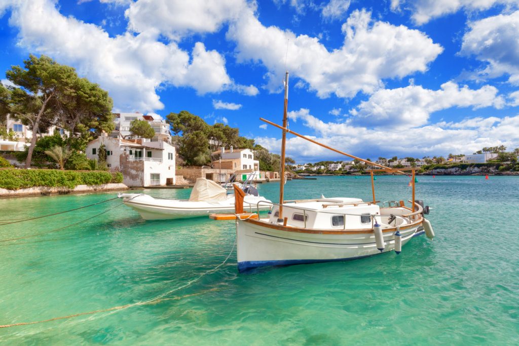 Traditionelles Fischerboot im türkisblauen Wasser von Cala d’Or auf Mallorca, umgeben von weißen Häusern und grüner Vegetation unter blauem Himmel mit Wolken.