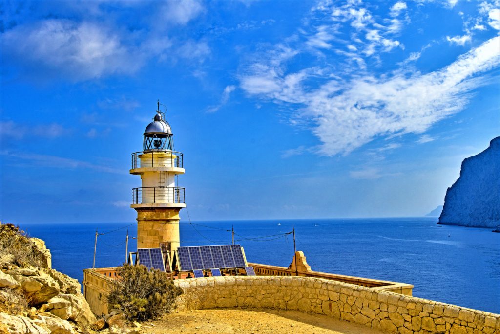Leuchtturm mit Solarpanels auf der Insel Dragonera bei Mallorca, vor blauem Himmel und dem Mittelmeer im Hintergrund.