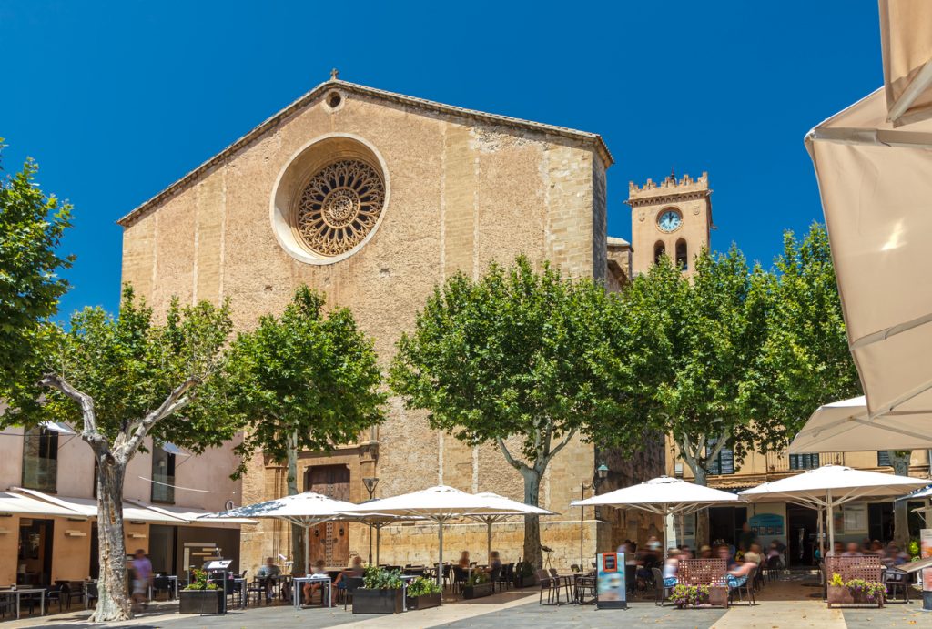 Placa Major in Alcudia auf Mallorca mit Kirche, Bäumen und Straßencafés an einem sonnigen Tag.