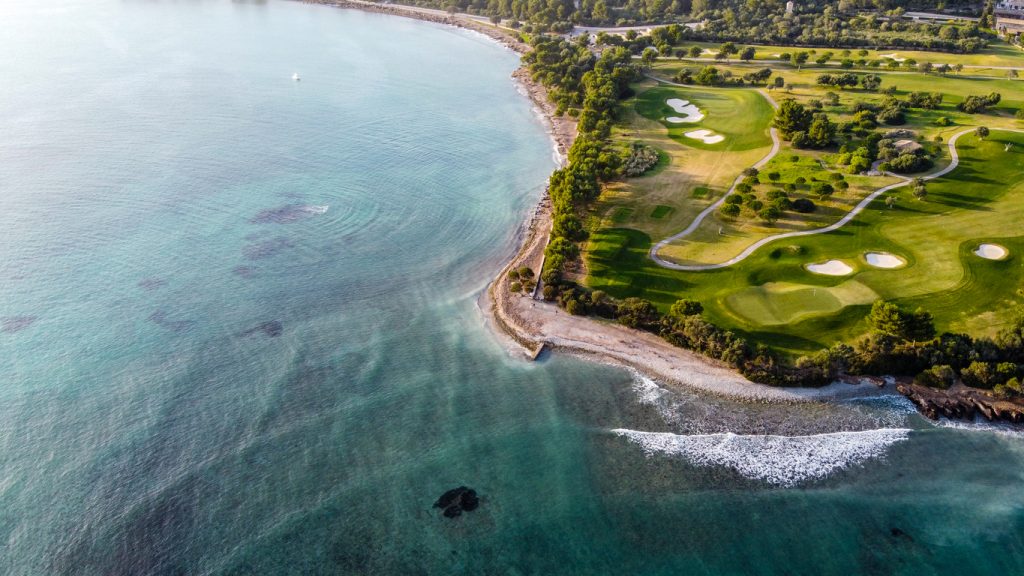 Luftaufnahme eines Golfplatzes auf Mallorca direkt an der Küste mit türkisblauem Meer, Sandbunkern und gepflegten Fairways.
