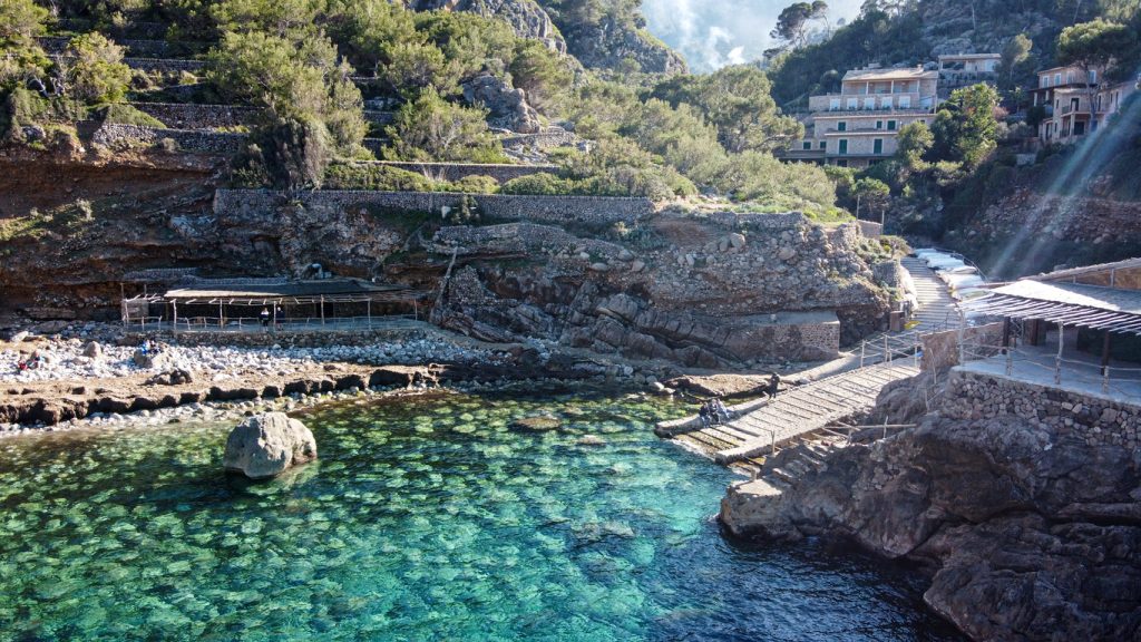 Kiesbucht Cala Deià mit türkisfarbenem, kristallklarem Wasser, Felsen und rustikalen Bootsgaragen im Tramuntana-Gebirge