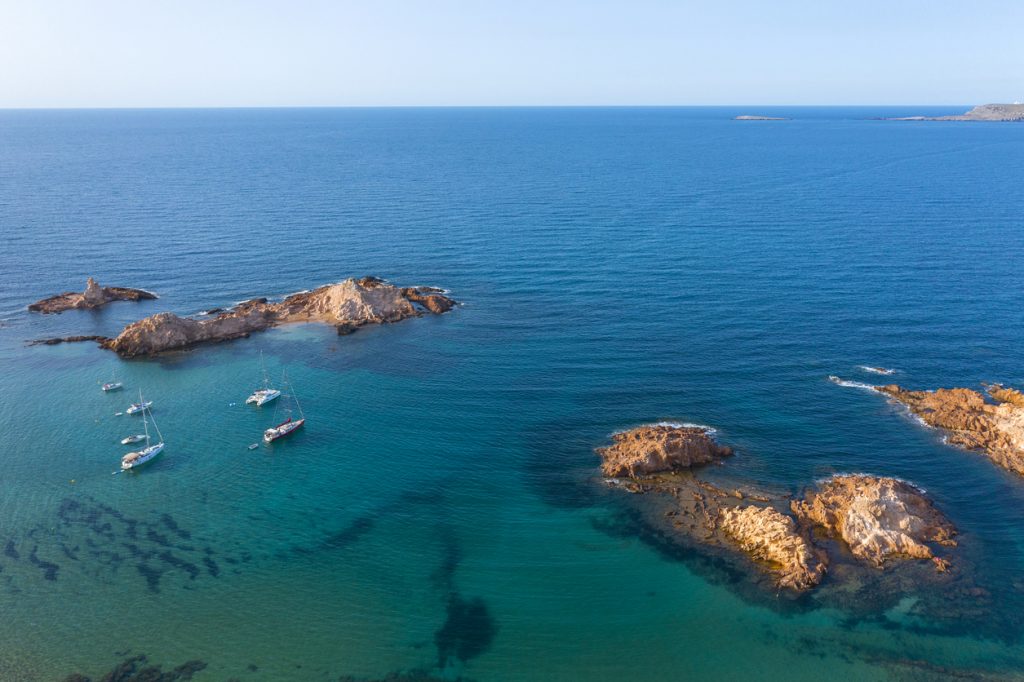 Drohnenaufnahme von Segelbooten vor den Felseninseln in der türkisblauen Bucht Cala Pregonda auf Menorca