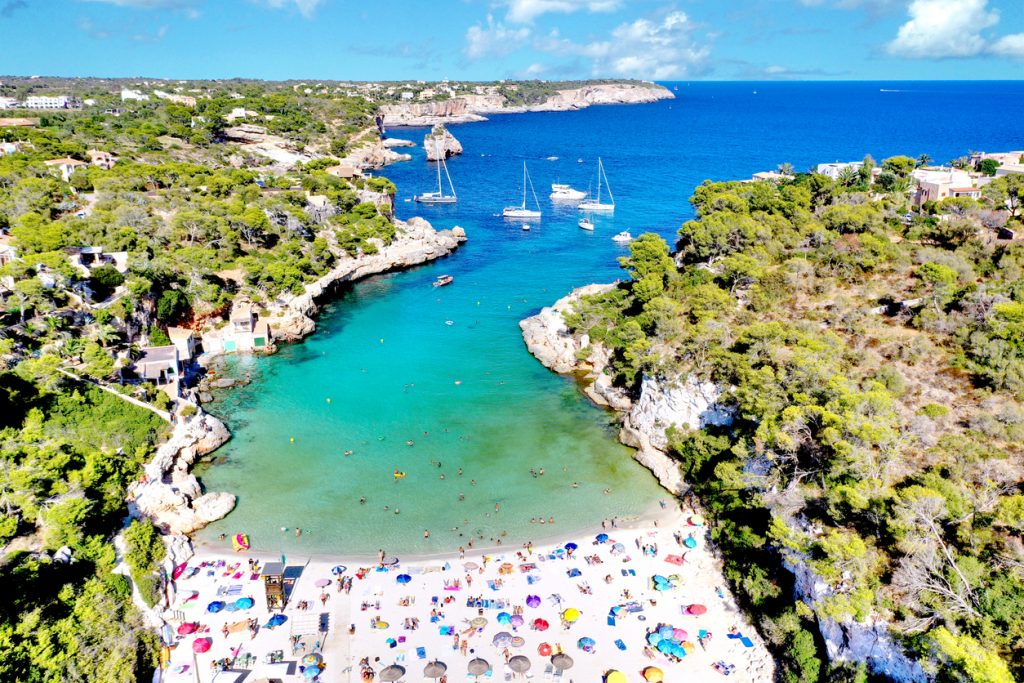 Luftaufnahme der Cala Llombards auf Mallorca mit feinem Sandstrand, bunten Sonnenschirmen, türkisblauem Wasser und Segelbooten in der Bucht.