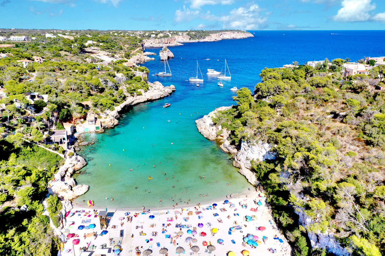 Luftaufnahme der Cala Llombards auf Mallorca mit feinem Sandstrand, bunten Sonnenschirmen, türkisblauem Wasser und Segelbooten in der Bucht.