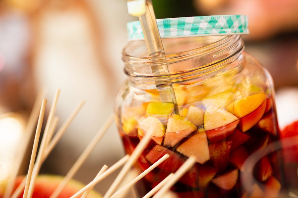 Großes Glasgefäß mit Sangria, gefüllt mit frischen Obststücken wie Pfirsichen und Äpfeln, dazu ein Schöpflöffel und Holzspieße im Vordergrund.