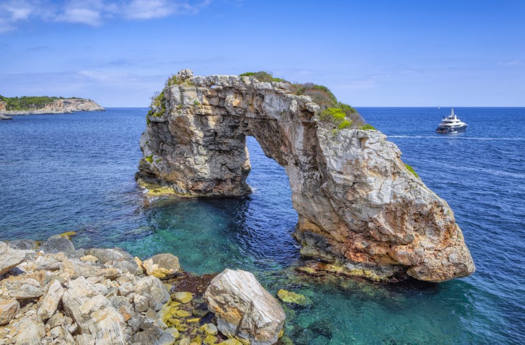 Natürlicher Felsbogen Es Pontàs an der Küste bei Cala Llombards auf Mallorca, umgeben von türkisblauem Meer, mit einem Boot im Hintergrund.