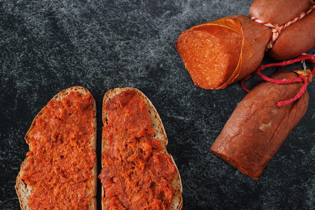 Zwei Scheiben Brot mit Sobrasada bestrichen, daneben ganze Würste der mallorquinischen Paprikawurst.
