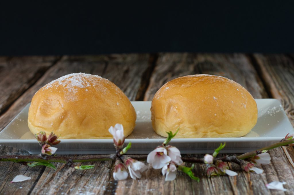 Zwei frische Coca de Patata auf einem rechteckigen weißen Teller, dekoriert mit Mandelblütenzweigen, auf rustikalem Holztisch.