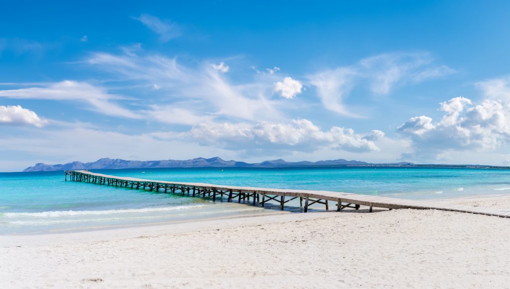Weißer Sandstrand von Alcudia auf Mallorca mit langem Holzsteg, türkisfarbenem Wasser und Bergen im Hintergrund.
