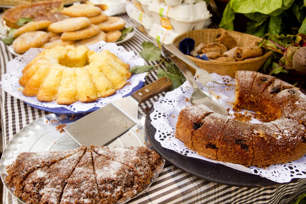 Verschiedene hausgemachte Kuchen auf Tellern mit Zuckerglasur, präsentiert auf einem Wochenmarktstand auf Mallorca, daneben Gebäck, Eier und frisches Gemüse.