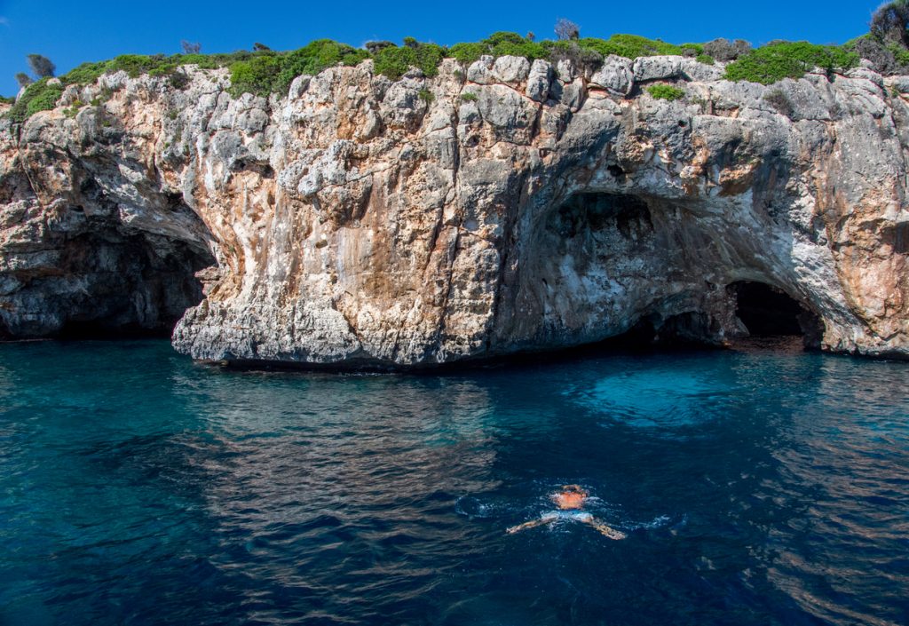 Person schwimmt vor steiler Felswand mit Meereshöhlen an der Küste von Mallorca