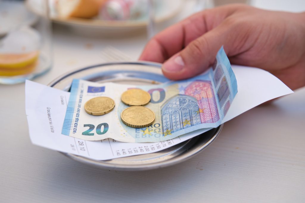 Nahaufnahme von einem 20-Euro-Schein mit Euromünzen auf einem kleinen Metallteller zusammen mit einem Kassenbon und einer Hand
