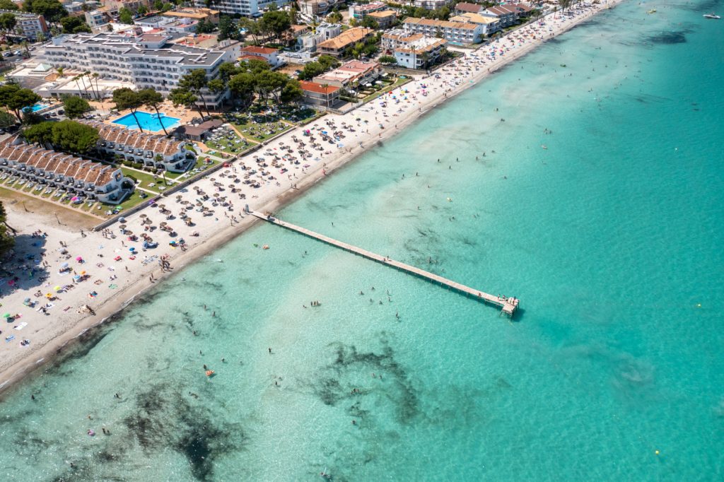 Luftaufnahme des Strandes von Alcudia auf Mallorca mit Hotelanlagen, Liegen, Sonnenschirmen und türkisblauem Meer.