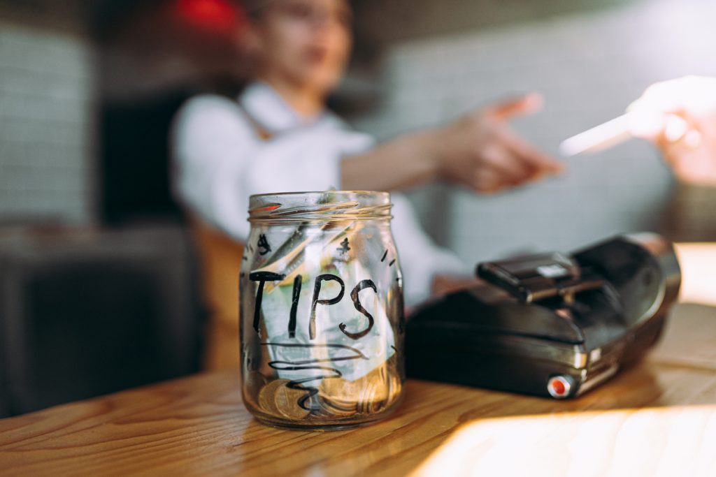 Ein Glas mit der Aufschrift "TIPS", gefüllt mit Geldscheinen und Münzen, steht auf einem Tresen neben einem Kartenterminal. Im Hintergrund ist unscharf eine Servicekraft zu sehen, die einem Gast die Quittung reicht.