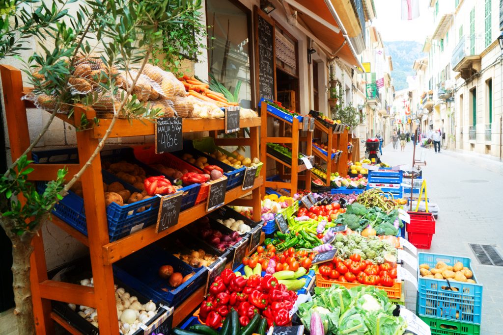 Straßenmarkt in Sóller, Mallorca, mit einem üppigen Gemüsestand voller Paprika, Tomaten, Zucchini, Zwiebeln und anderer frischer Produkte in Holzauslagen entlang einer charmanten Altstadtgasse.