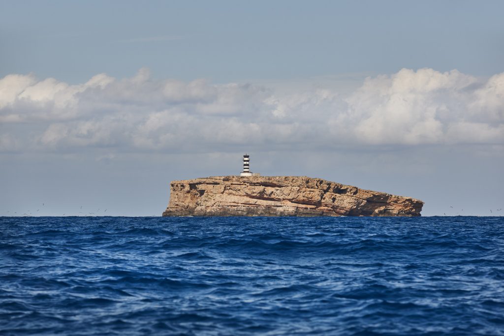 Flache Felseninsel mit schwarz-weiß gestreiftem Leuchtturm im Mittelmeer vor bewölktem Himmel