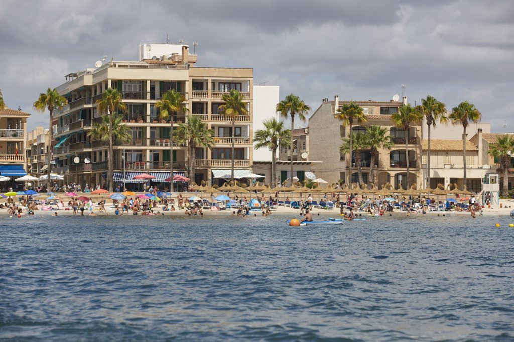 Menschen genießen den Sommer am Strand von Colònia de Sant Jordi auf Mallorca vor Hotels und Palmen