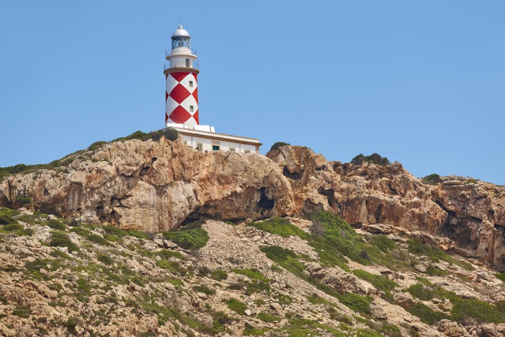 Leuchtturm mit rot-weißem Rautenmuster auf felsigem Hügel der Insel Cabrera bei klarem Himmel