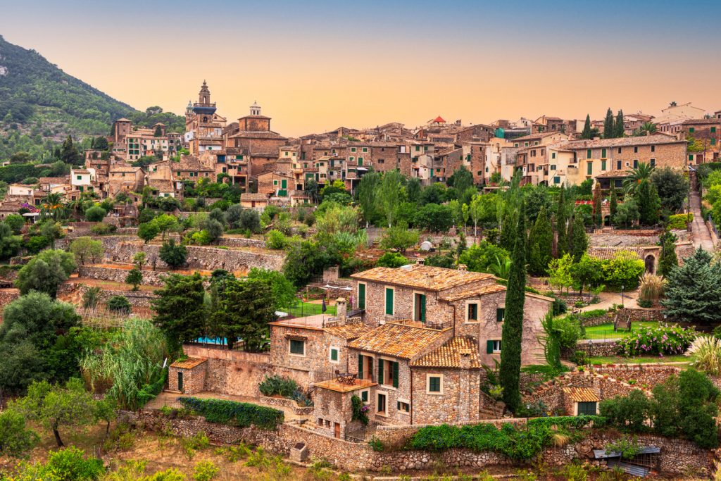 Blick auf das malerische Bergdorf Valldemossa auf Mallorca mit traditionellen Steinhäusern, grünen Terrassengärten und der umliegenden Tramuntana-Bergkulisse bei Sonnenuntergang.