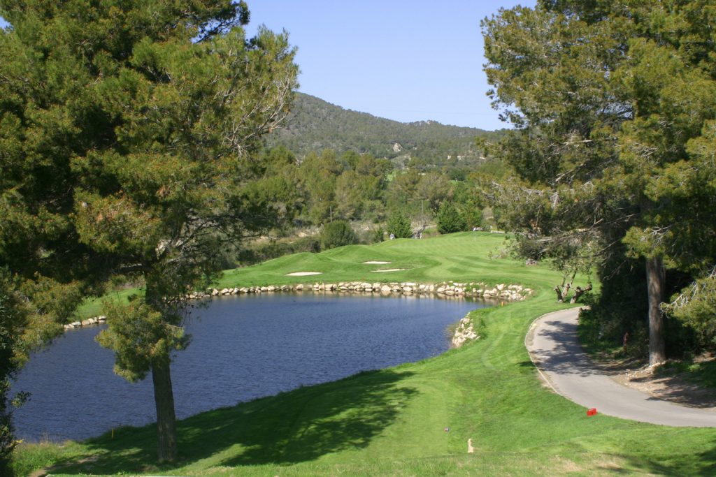 Blick auf einen gepflegten Golfplatz auf Mallorca mit sattem Grün, Bunkern und einem See, umgeben von Bäumen und Hügeln im Hintergrund.