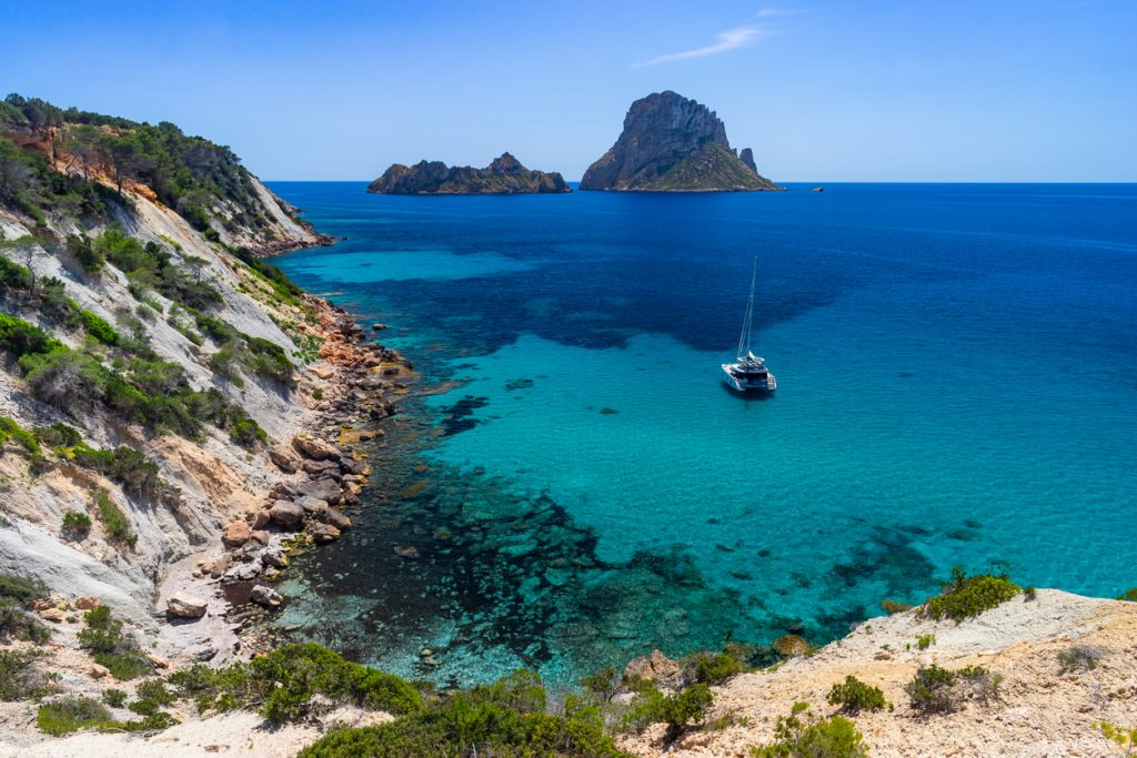 Blick auf die Felseninsel Es Vedrà vor Ibiza mit türkisblauem Wasser und Segelboot bei Cala d’Hort.