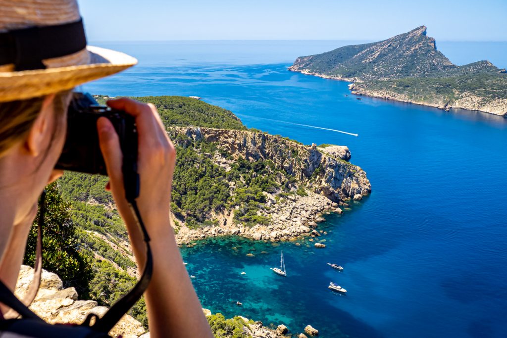 Fotograf mit Strohhut fotografiert die Insel Dragonera vor der Küste Mallorcas, mit türkisblauem Wasser, Segelbooten und felsiger Küstenlandschaft.