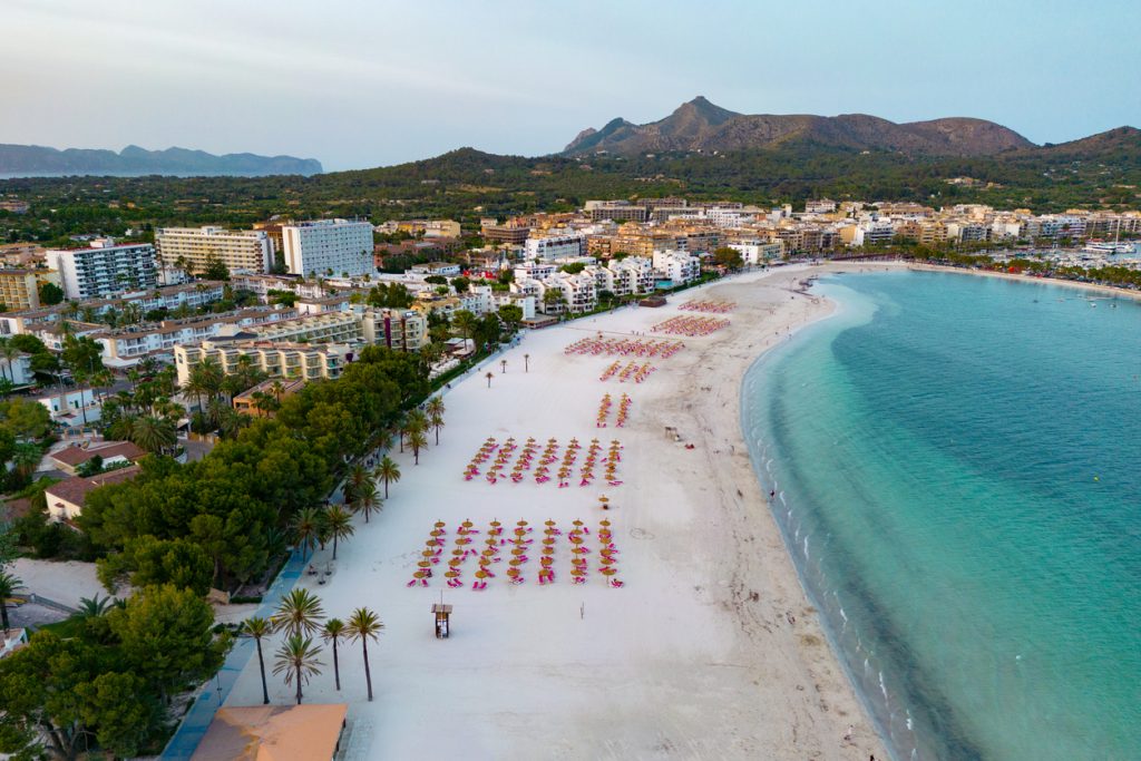 Luftaufnahme des Strandes von Alcudia mit weißen Sandstränden, Sonnenschirmen, türkisfarbenem Wasser und der Stadt im Hintergrund.