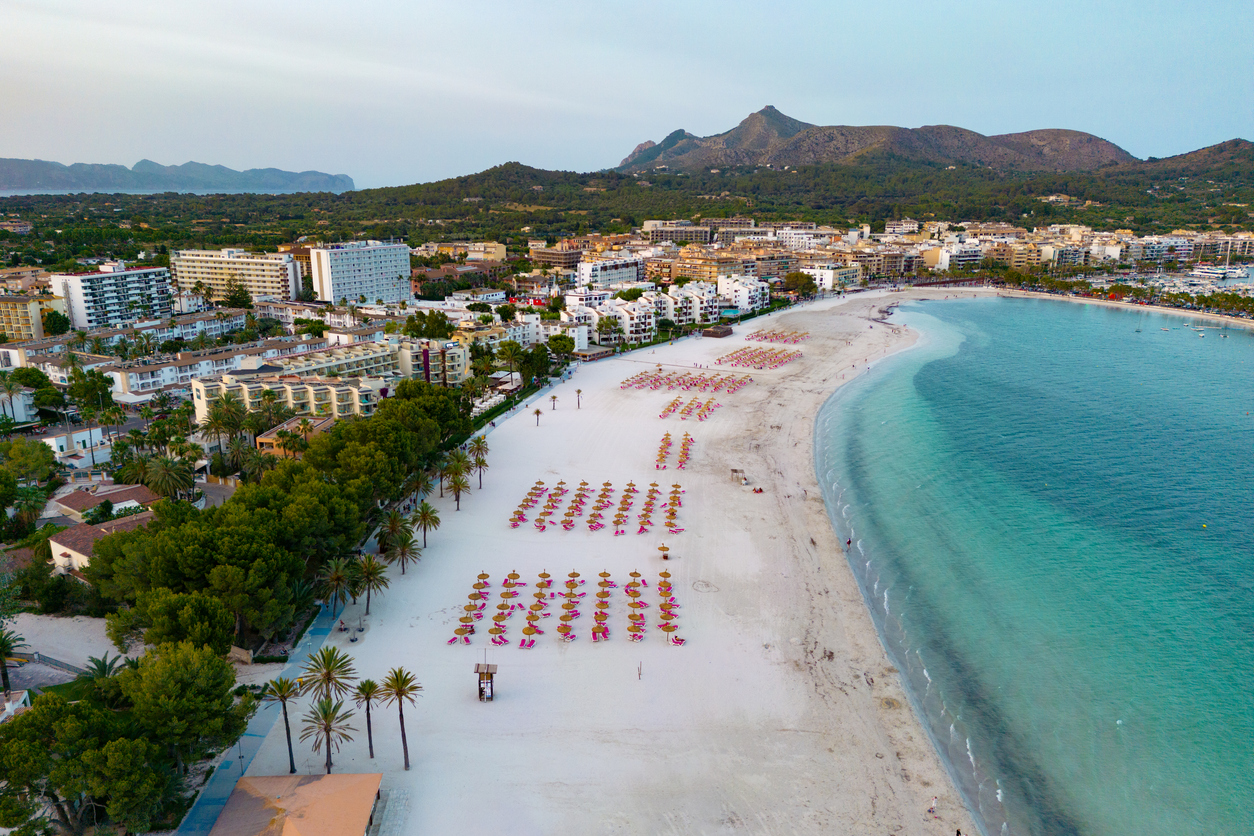 Luftaufnahme des Strandes von Alcudia mit weißen Sandstränden, Sonnenschirmen, türkisfarbenem Wasser und der Stadt im Hintergrund.
