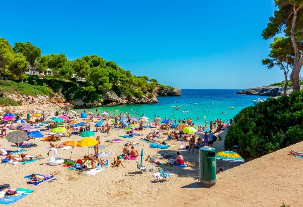 Blick auf den belebten Strand von Cala d’Or auf Mallorca, voller bunter Sonnenschirme und Urlauber am türkisblauen Meer.