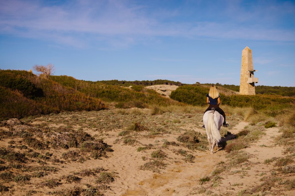 Blonde Reiterin auf weißem Pferd reitet durch Dünenlandschaft auf Mallorca in Richtung eines alten Wachturms