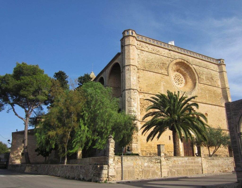 Historische Kirche in Petra, Mallorca, aus hellem Sandstein mit Rosettenfenster, flankiert von Palmen und Bäumen vor blauem Himmel.