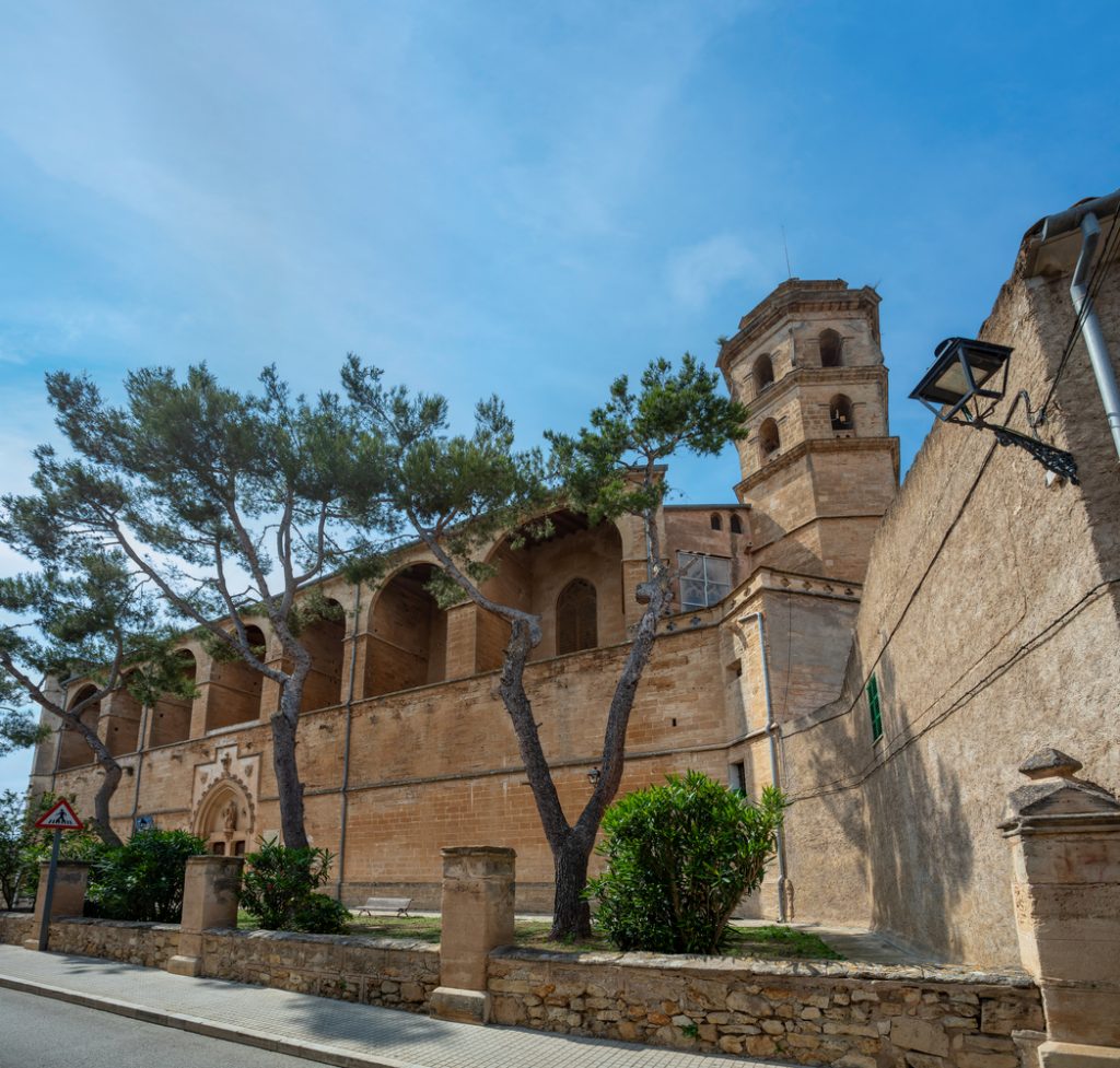 Historische Kirche in Petra, Mallorca, aus Sandstein mit markantem Glockenturm, flankiert von Pinienbäumen und einer Steinmauer unter blauem Himmel.