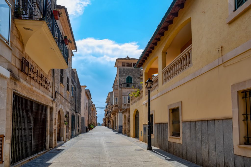 Leere Straße in Petra auf Mallorca, gesäumt von historischen Sandsteinhäusern und modernen Gebäuden, unter blauem Himmel mit weißen Wolken.