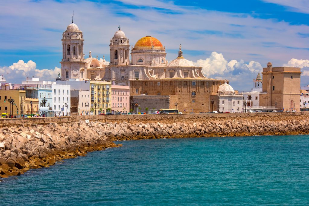 Die Kathedrale von Cádiz an der Uferpromenade mit bunten Häusern und dem Atlantik im Vordergrund.