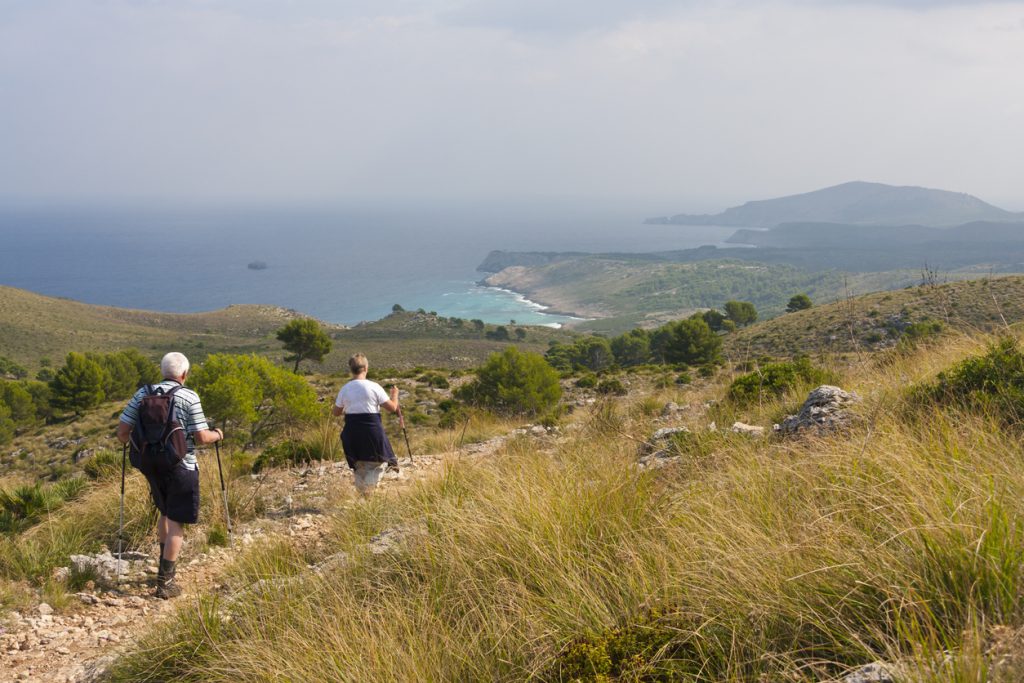 Zwei Wanderer mit Trekkingstöcken gehen einen Pfad hinunter und genießen den Meerblick auf Mallorca im herbstlichen Sonnenlicht.