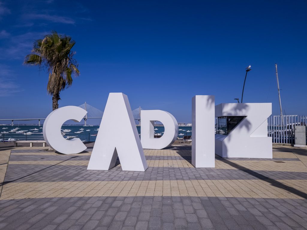 Große weiße Buchstaben formen den Schriftzug Cádiz am Hafen mit Meer, Booten und Brücke im Hintergrund.