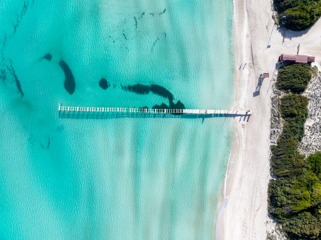 Luftaufnahme des Strandes von Alcudia mit einem langen Holzsteg, türkisfarbenem Wasser, Badenden und Hotels im Hintergrund.