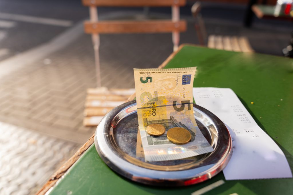 Nahaufnahme eines Trinkgelds mit einem 5-Euro-Schein und zwei Euromünzen auf einem Kellnertablett neben einem Kassenbon