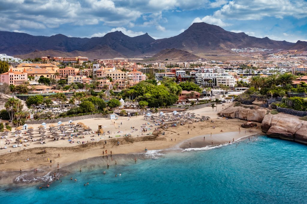 Strand Playa del Duque in Costa Adeje mit goldenem Sand, türkisblauem Wasser und luxuriösen Hotels im Hintergrund.
