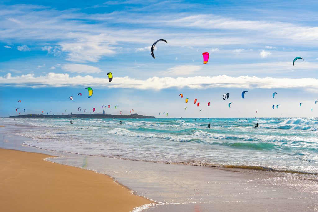 Bunte Kitesegel über dem Meer am Strand von Cádiz mit Surfern in den Wellen.