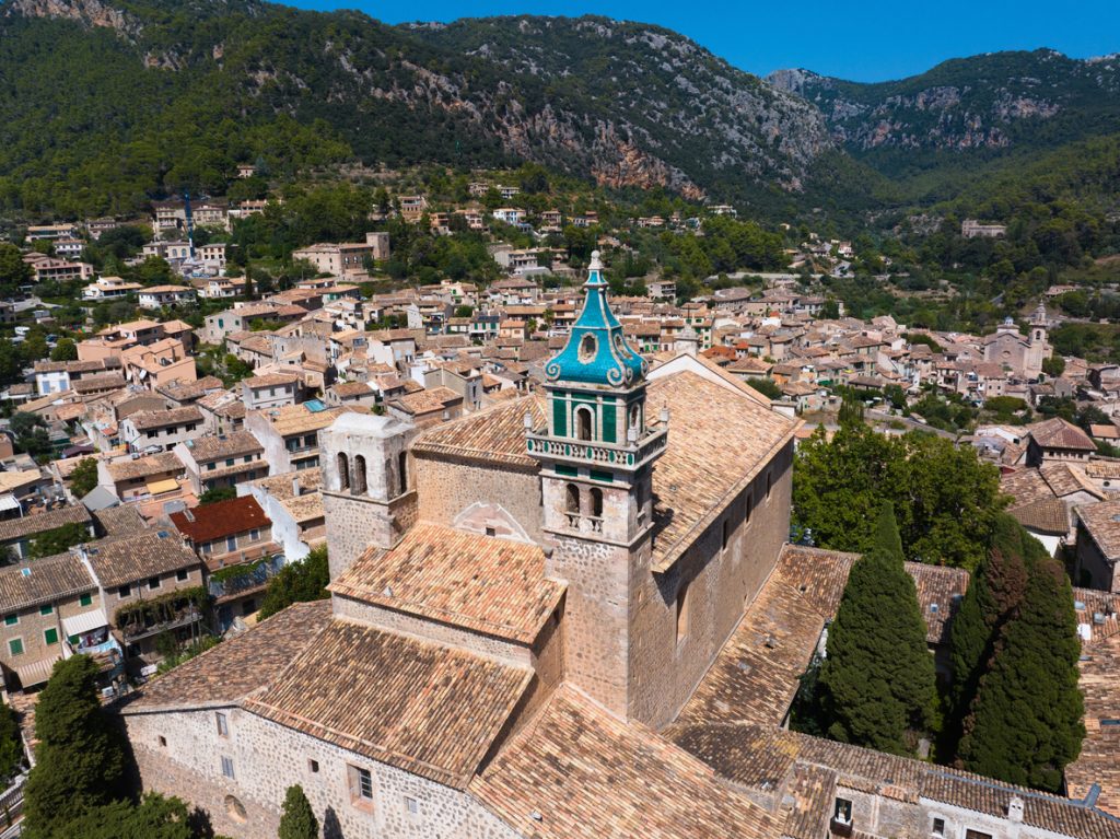 Luftaufnahme des Kartäuserklosters in Valldemossa auf Mallorca mit markantem grünen Glockenturm, umgeben von traditionellen Steinhäusern und den Bergen der Serra de Tramuntana.