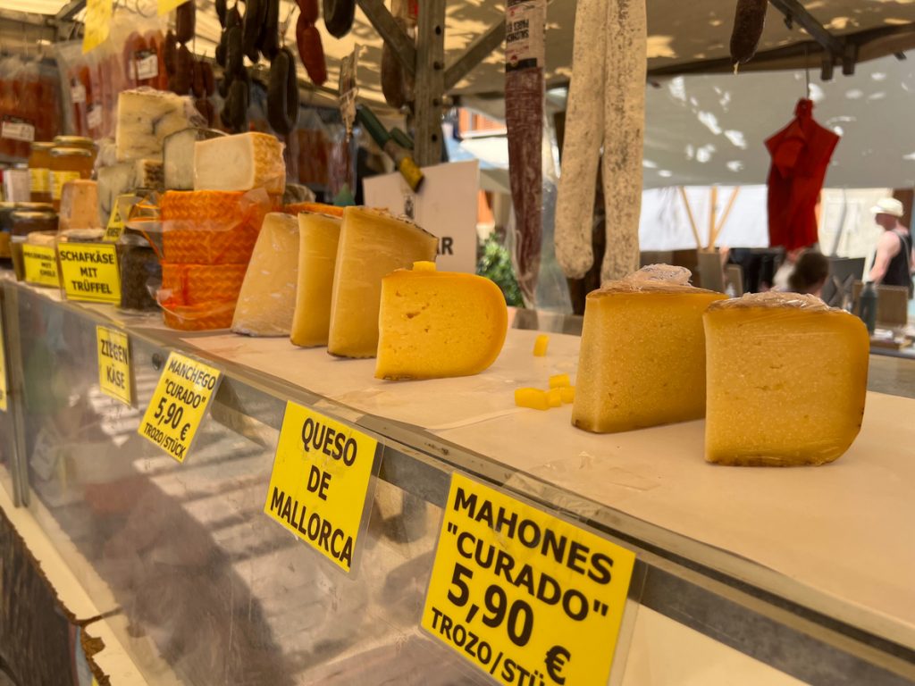 Verschiedene Sorten Käse, darunter Queso de Mallorca, Mahonés und Manchego, auf einem Marktstand auf Mallorca, im Hintergrund Wurstwaren und andere Spezialitäten.