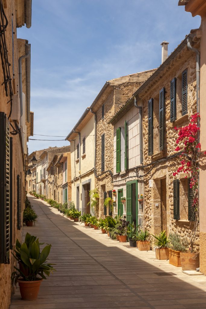 Schmale historische Gasse in Alcudia mit Naturstein-Häusern, grünen Fensterläden und Blumentöpfen.