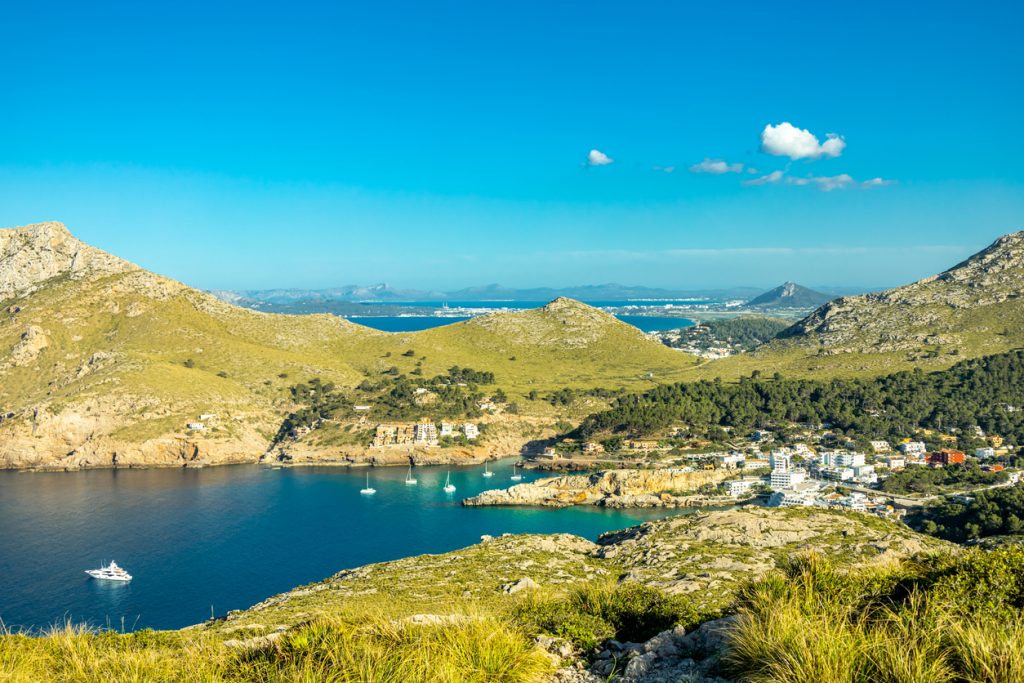 Blick auf Cala Sant Vicenç mit Küstenort, umliegenden Bergen und mehreren Yachten in der Bucht